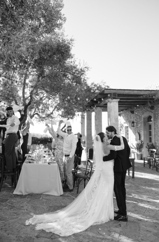 A bride and groom share a kiss outdoors beside a long dining table, while guests stand and celebrate, some with raised arms. The scene is joyful and festive, set against a stone building and trees.