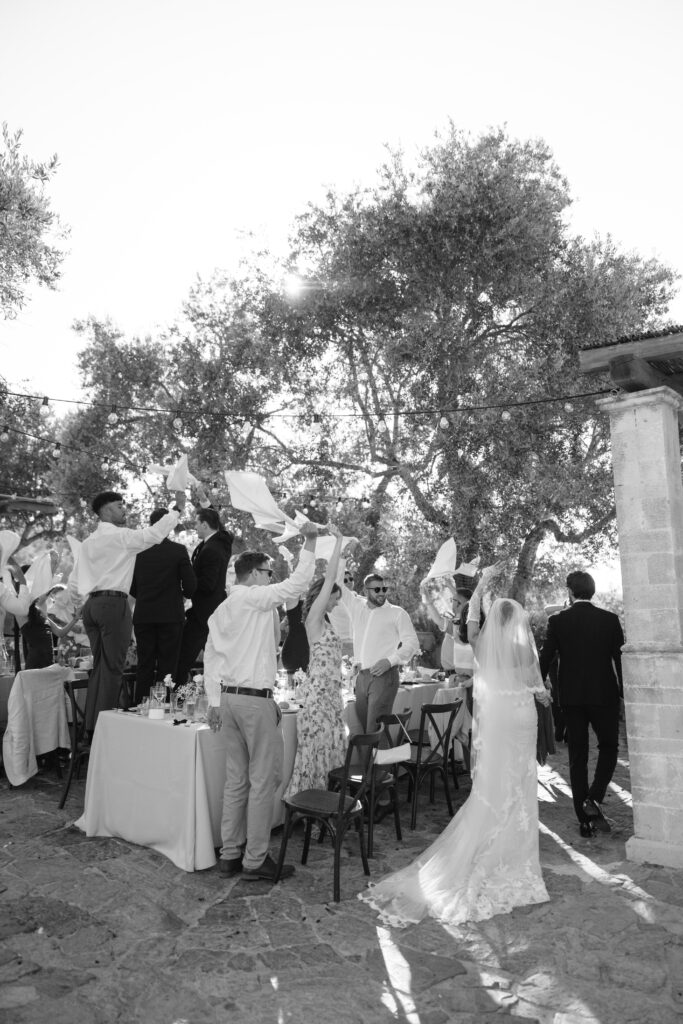 A bride and groom walk past outdoor wedding guests who stand on chairs and wave napkins joyfully. The scene is festive, with sunlight filtering through large trees in the background.