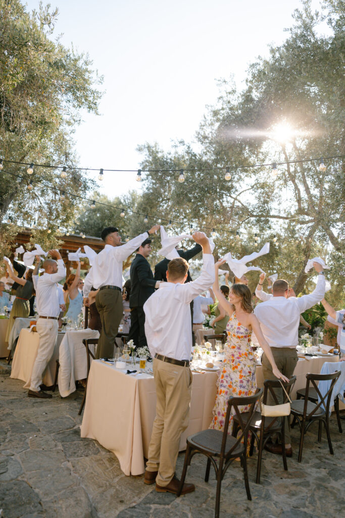 A group of people at an outdoor event stand and wave napkins in the air, some standing on chairs or tables. The scene is festive, with sunlight filtering through trees and string lights overhead.