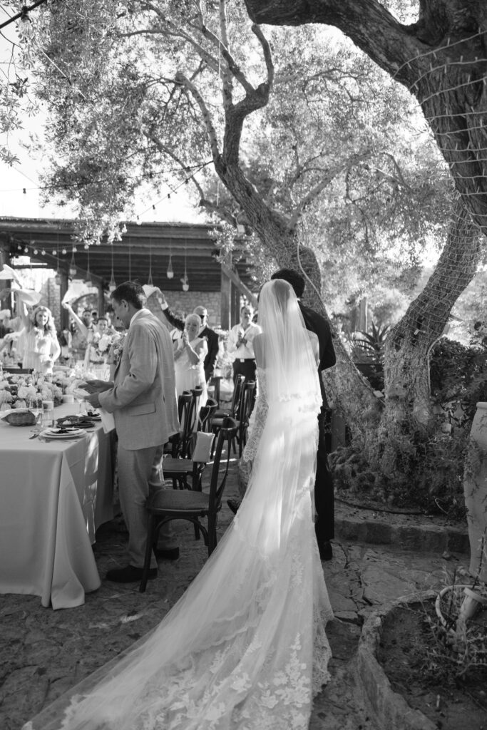 A bride in a long lace gown and veil walks towards a group of seated and standing guests at an outdoor wedding reception under trees, with sunlight filtering through the leaves.