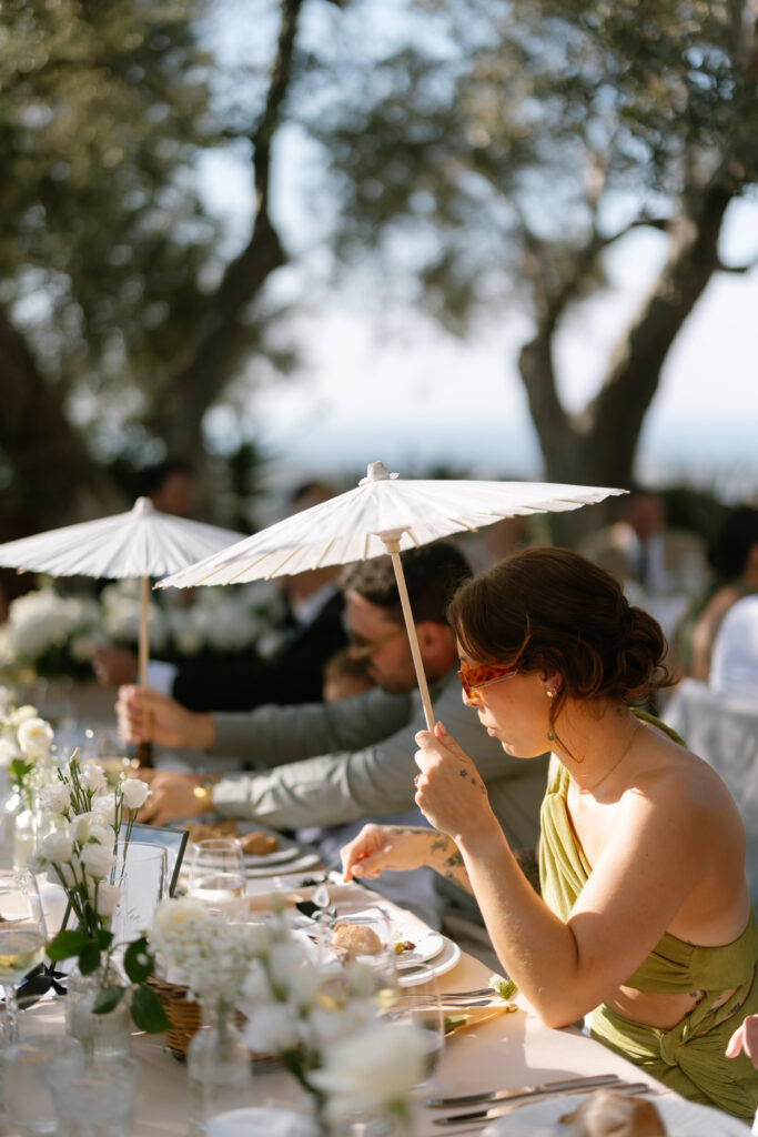 A woman in a green dress holds a white parasol while sitting at an outdoor table set for a meal. Others seated nearby also hold parasols. The setting is sunny, with trees and floral centerpieces visible.