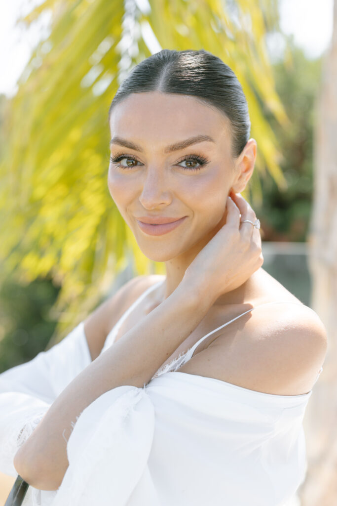 A woman with dark hair pulled back and glowing makeup smiles gently, touching her neck with one hand. She wears a white off-shoulder top and stands outdoors with greenery and sunlight in the background.