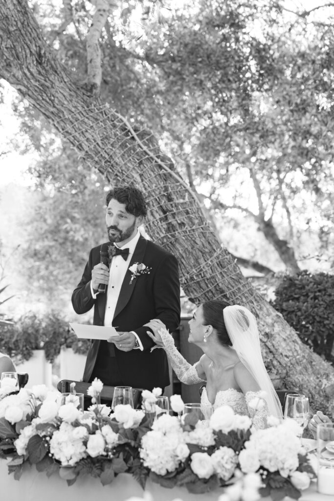 A groom in a tuxedo gives a speech with a microphone while a bride in a wedding dress and veil sits beside him, smiling and touching his hand, at a flower-adorned table outdoors.