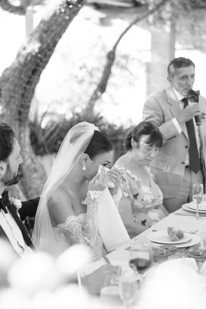 A bride in a wedding dress wipes tears with a napkin during a speech. A man in a suit speaks into a microphone, while others sit at the table set with food and drinks. The scene is outdoors in natural light.