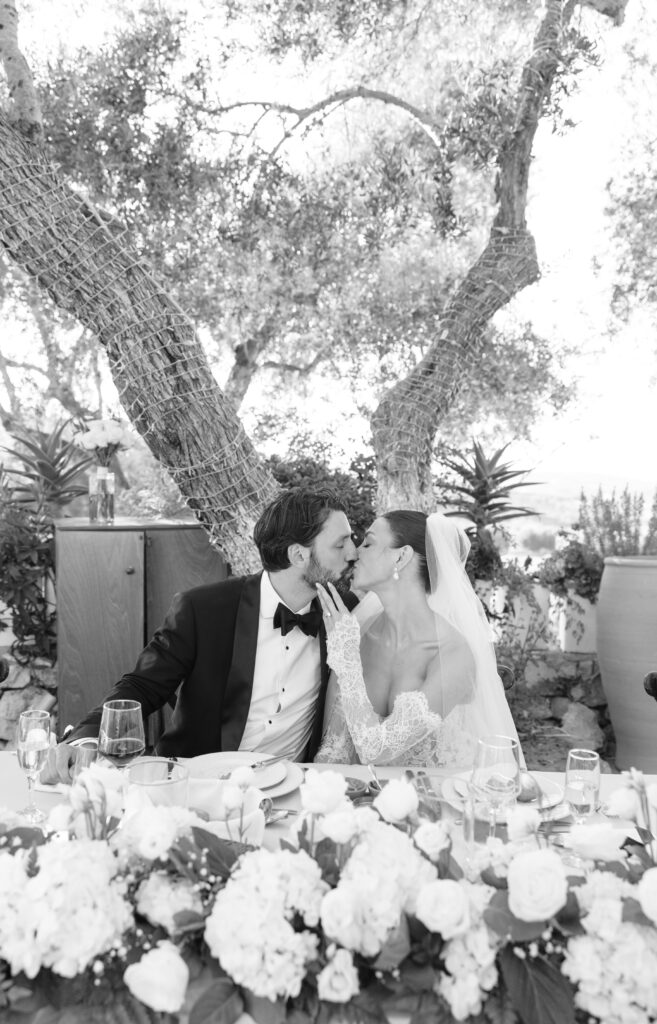 A bride and groom sit at a decorated table outdoors, sharing a kiss. The bride wears a veil and lace dress, while the groom is in a tuxedo. Lush floral arrangements and trees surround the couple.
