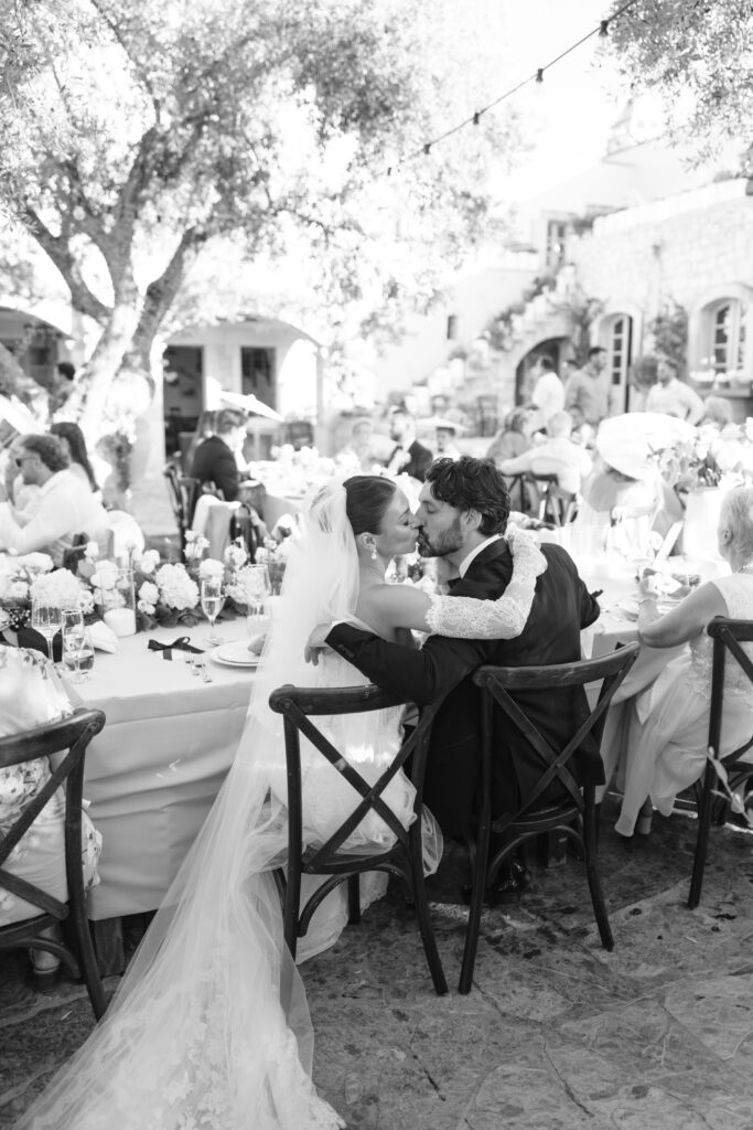 A bride and groom sit closely together at an outdoor wedding reception, sharing an intimate moment amid guests seated at decorated tables under string lights.