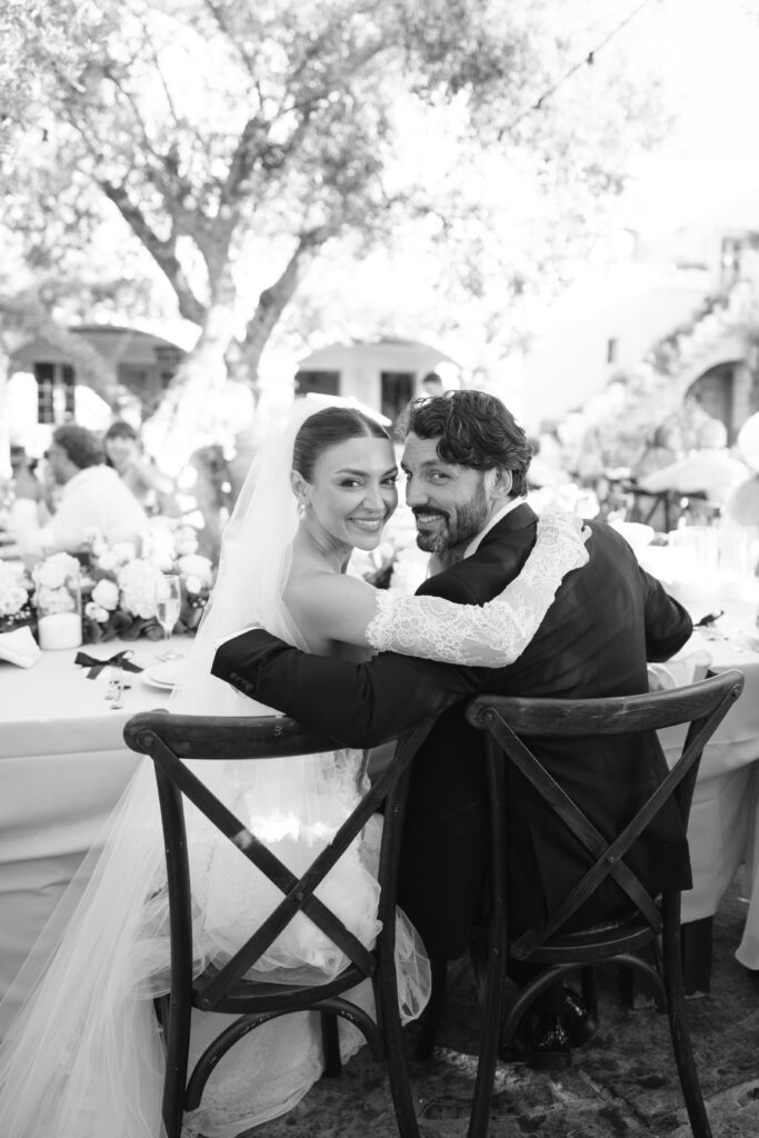 A bride and groom sit closely together on chairs at an outdoor wedding reception, smiling and looking back at the camera; the bride wears a veil and dress, the groom is in a suit, and guests are visible in the background.
