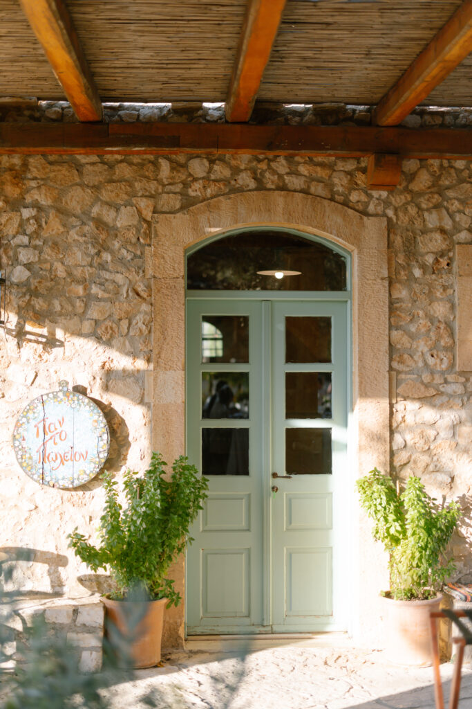 A light blue wooden door with glass panels set in a rustic stone wall, flanked by two potted green plants, with a round sign featuring Greek text on the left side of the entrance. Sunlight casts a warm glow.