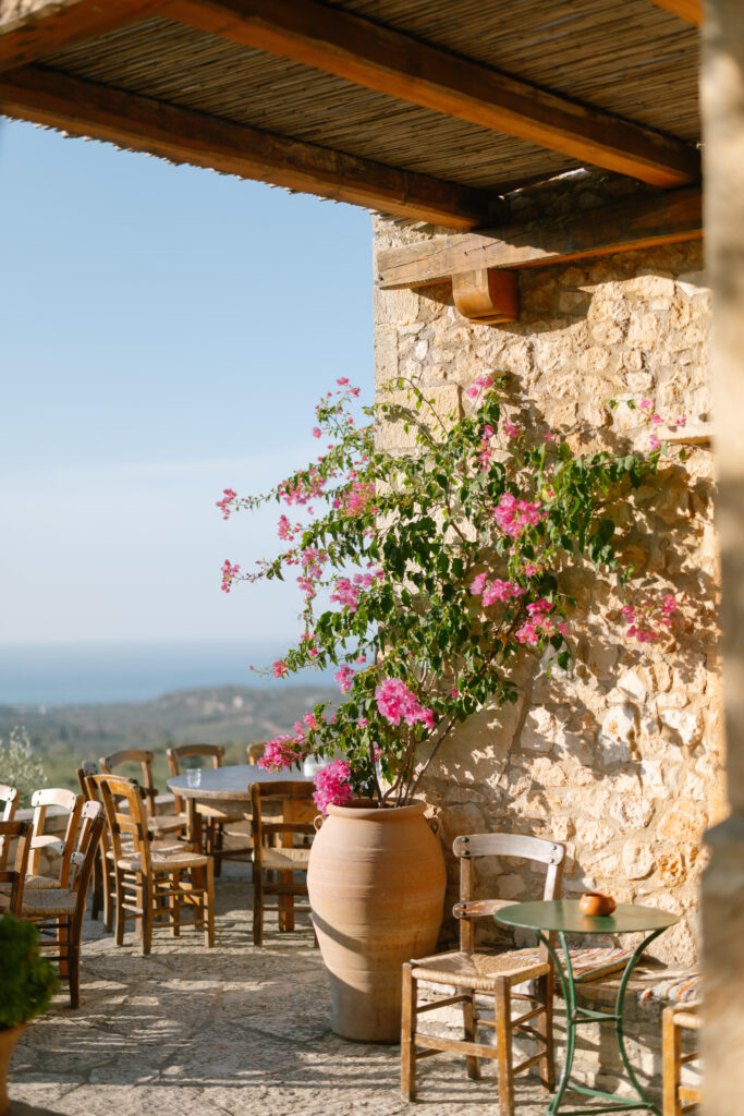 A sunlit stone patio with wooden tables and chairs overlooks a distant blue sea. Pink bougainvillea climbs a wall beside a large clay pot, creating a peaceful Mediterranean atmosphere.