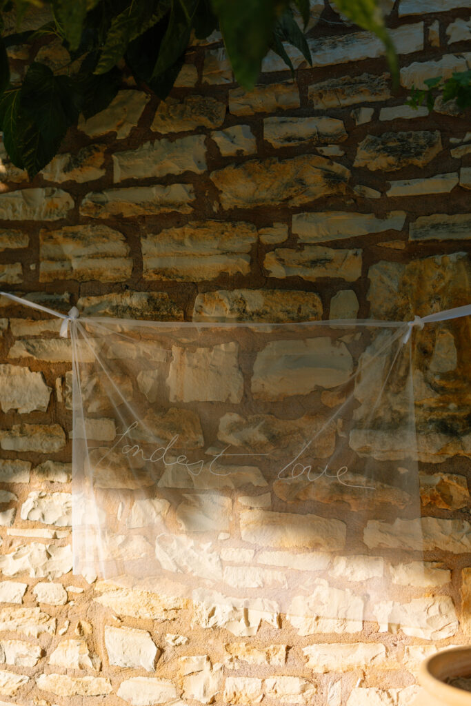 A sheer fabric banner with the handwritten words kindest love hangs between two cords against a sunlit stone wall, with green leaves partially visible at the top.