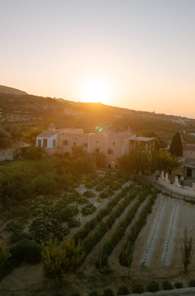 Aerial view of a house surrounded by gardens and farmland at sunrise, with a golden glow illuminating the landscape and hills in the background.