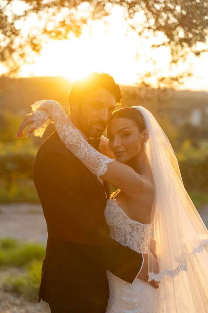 A bride and groom embrace outdoors at sunset, bathed in warm golden light. The bride wears a lace wedding dress and veil, while the groom wears a dark suit. Both look content and peaceful.