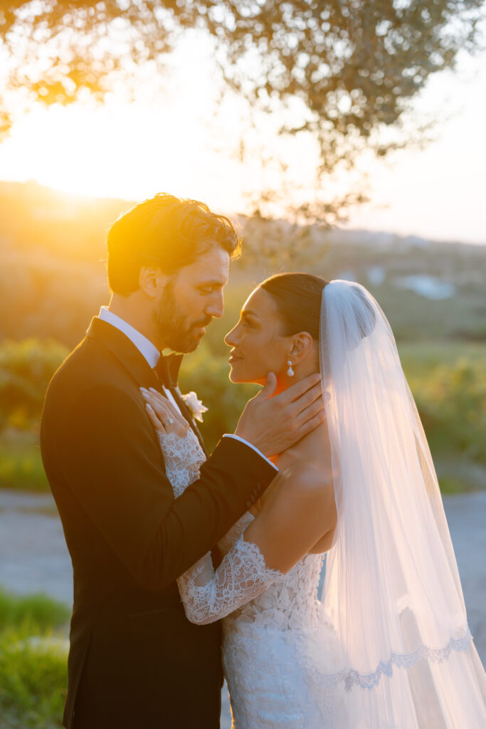 A bride and groom stand close together outdoors at sunset, gazing into each others eyes. The bride wears a white lace gown and veil, while the groom is in a black suit. Sunlight softly illuminates them from behind.