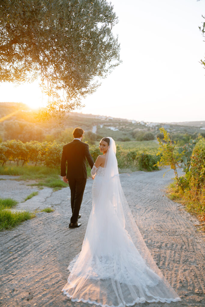 A bride in a white dress and veil looks back and smiles while holding hands with a groom in a suit as they walk down a sunlit path surrounded by greenery at sunset.