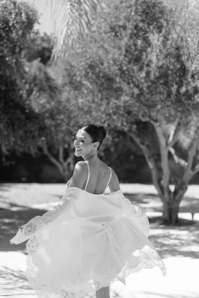 A woman with dark hair in a bun twirls outdoors, smiling over her shoulder. She wears a light, lace-trimmed robe, with trees and sunlight in the background. The image is in black and white.