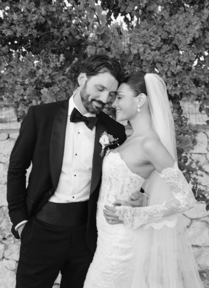A bride and groom stand close together outdoors, smiling and touching foreheads. The bride wears a lace wedding dress and veil, and the groom is in a tuxedo with a bow tie. Lush greenery and a stone wall are in the background.
