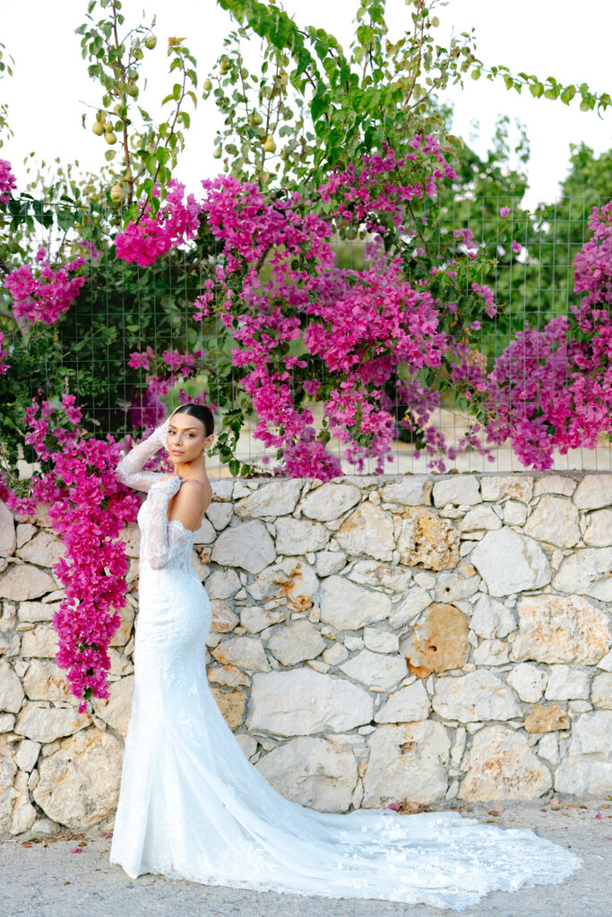 A bride in a long white lace gown poses beside a stone wall adorned with vibrant pink bougainvillea flowers and greenery under a clear sky.