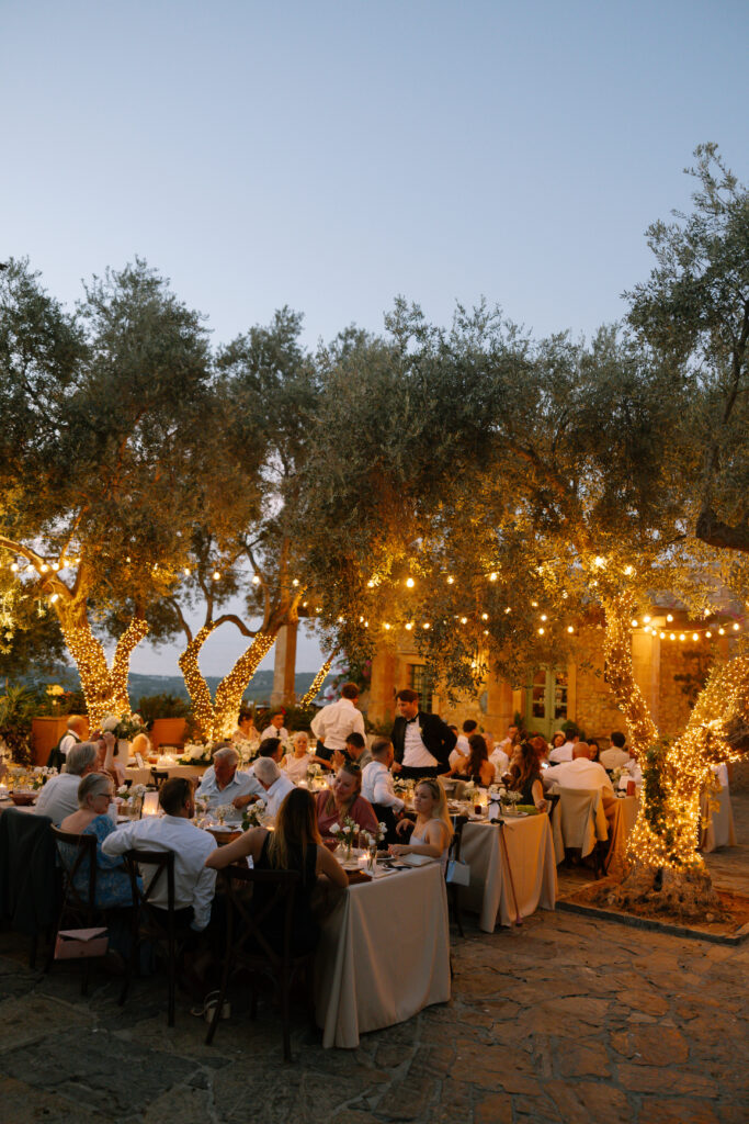 An outdoor evening event with guests dining at tables under large trees decorated with string lights and fairy lights, creating a warm, festive atmosphere. The setting is rustic and elegant, with a stone patio floor.