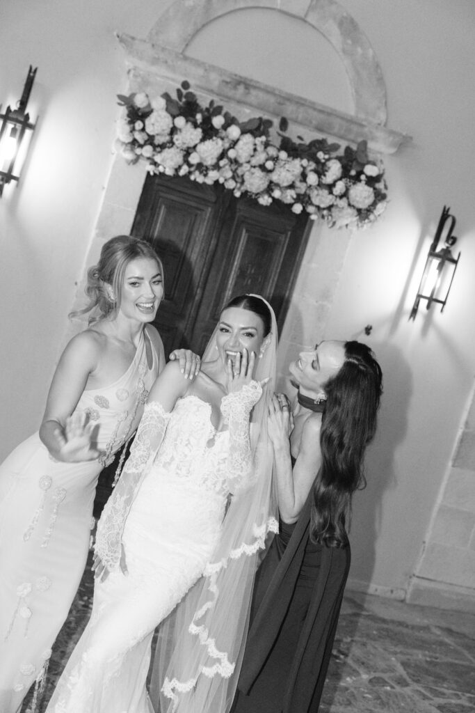 Three women in elegant dresses, including a bride in a lace gown and veil, stand smiling and laughing together in front of a flower-adorned door. The atmosphere is joyful and celebratory. The image is in black and white.