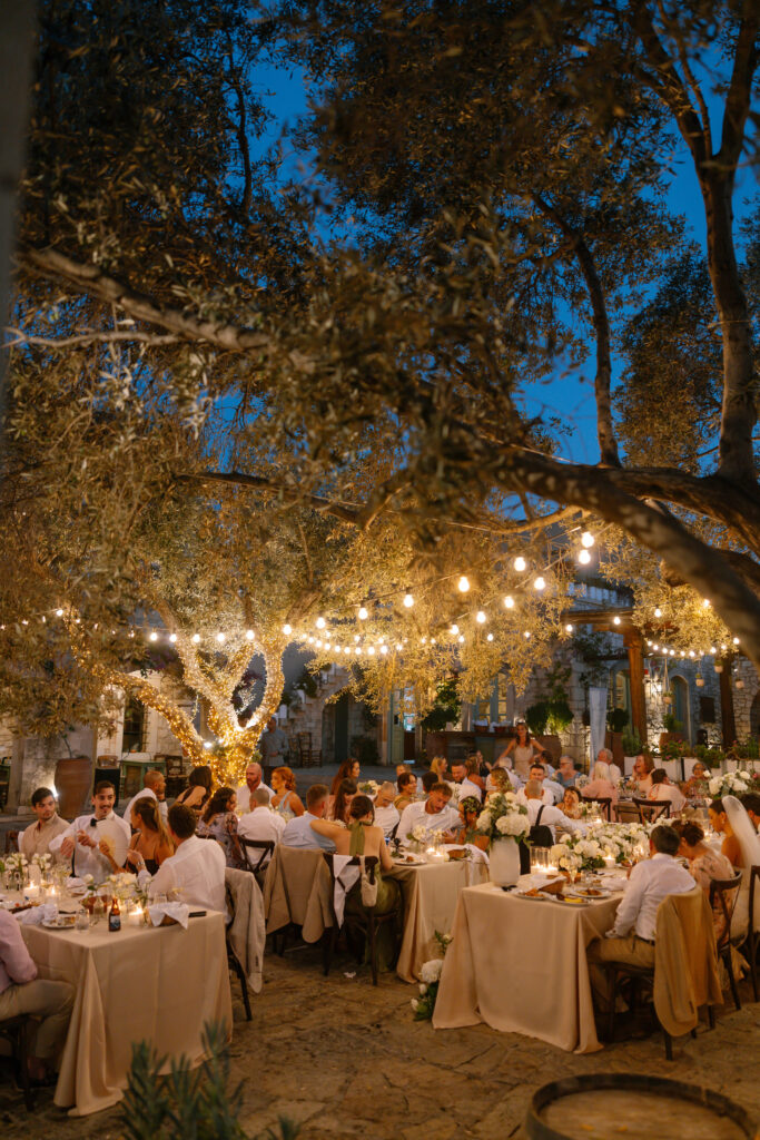 An outdoor evening event with guests seated at tables under trees decorated with string lights. The tables have cream tablecloths and floral centerpieces, creating a warm, festive atmosphere.