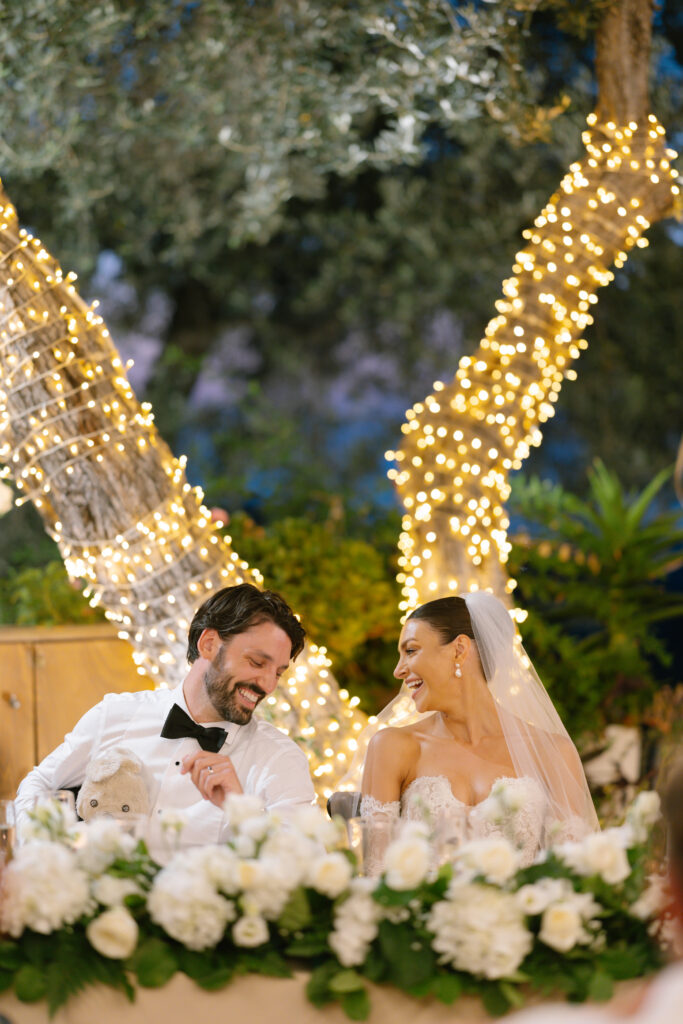 A bride and groom sit together, smiling joyfully at each other, in front of a table decorated with white flowers. Behind them, two trees wrapped in string lights create a romantic outdoor setting.
