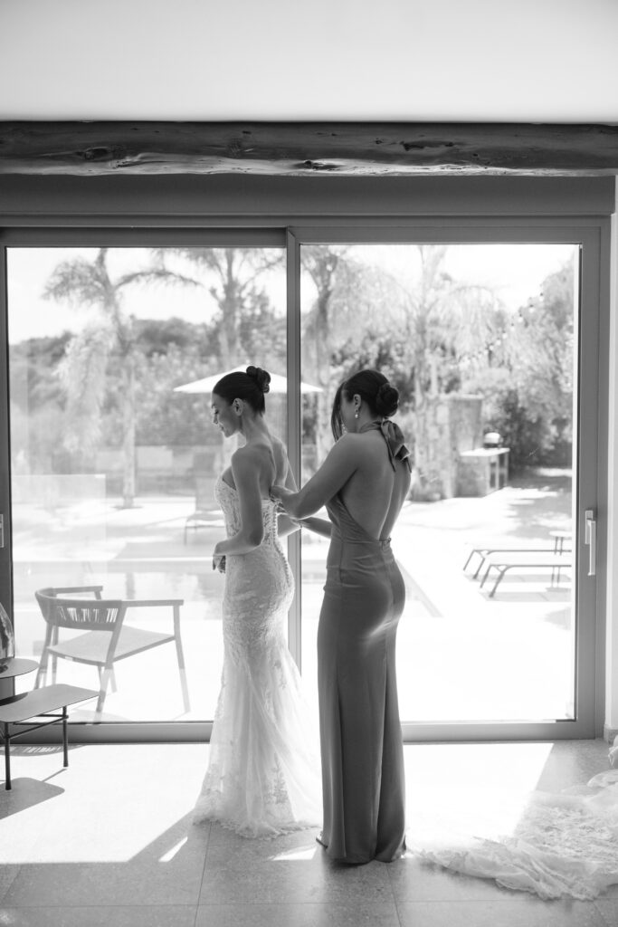 A woman in a formal dress helps a bride adjust her gown in front of large glass doors, with sunlight streaming in and a pool visible outside. The scene is elegant and serene.