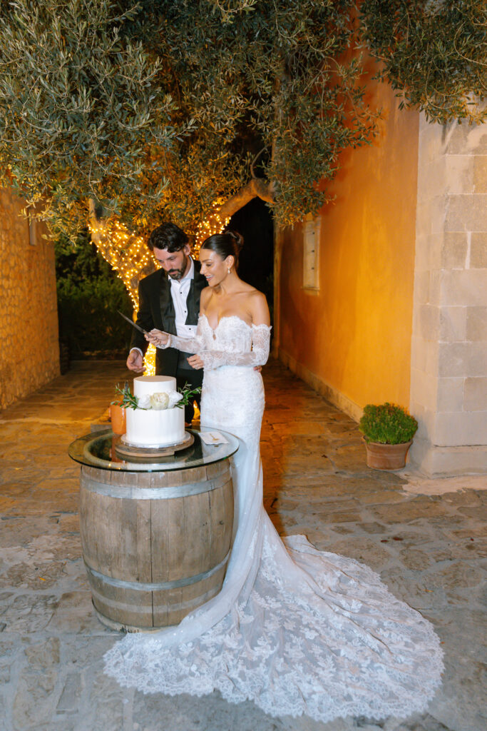 A bride and groom stand under a tree with string lights, cutting a white wedding cake on a wooden barrel outside a stone building at night. The bride wears a lace gown and the groom is in a black tuxedo.