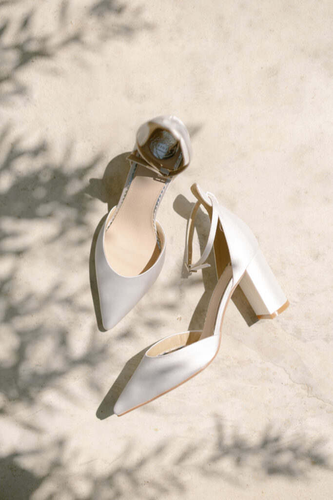 A pair of elegant white satin block-heeled shoes with pointed toes and ankle straps, placed on a light, sunlit surface with soft shadows of foliage.