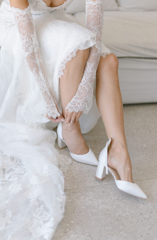 A bride in a lace wedding gown and matching lace gloves sits on a couch, adjusting a white block-heeled shoe on her foot, with her wedding dress spread around her.