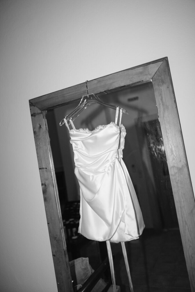 A short, white satin dress hangs on a clear hanger in front of a large wooden-framed mirror in a dimly lit room. The scene is in black and white.
