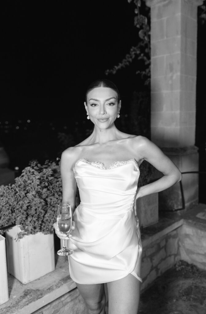 A woman in a strapless satin dress stands outdoors at night, holding a glass of wine and smiling. She has sleek hair pulled back and wears earrings, with potted plants and a stone wall in the background. The photo is black and white.