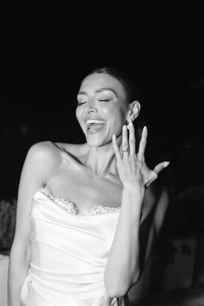 A woman in a satin dress smiles joyfully with her eyes closed, holding up her left hand to display a ring on her finger. The photo is in black and white with a dark background.