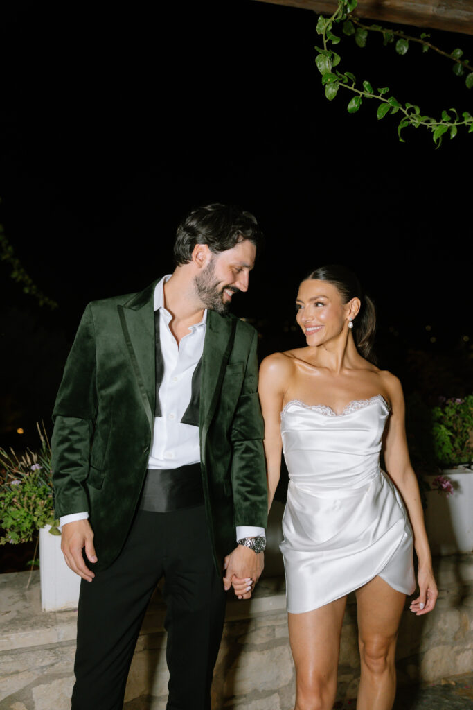 A man in a green velvet suit and a woman in a short white dress stand close together, holding hands and smiling at each other outdoors at night, with potted plants in the background.