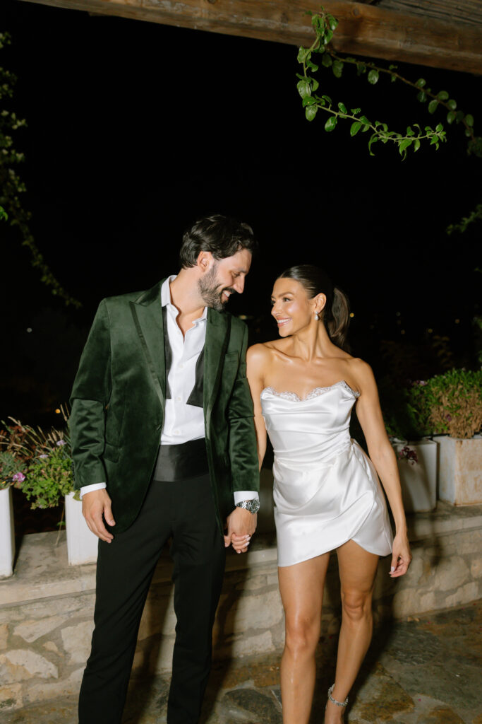 A man in a green velvet blazer and black pants holds hands with a woman in a short white satin dress. They smile at each other, standing outdoors at night with greenery and stone planters behind them.