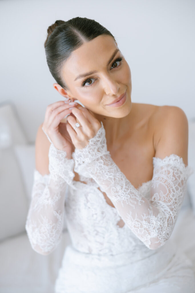A bride with dark hair in a sleek bun, wearing an off-the-shoulder lace wedding dress, smiles softly while putting on an earring. She is seated on a white couch with a bright, minimal background.