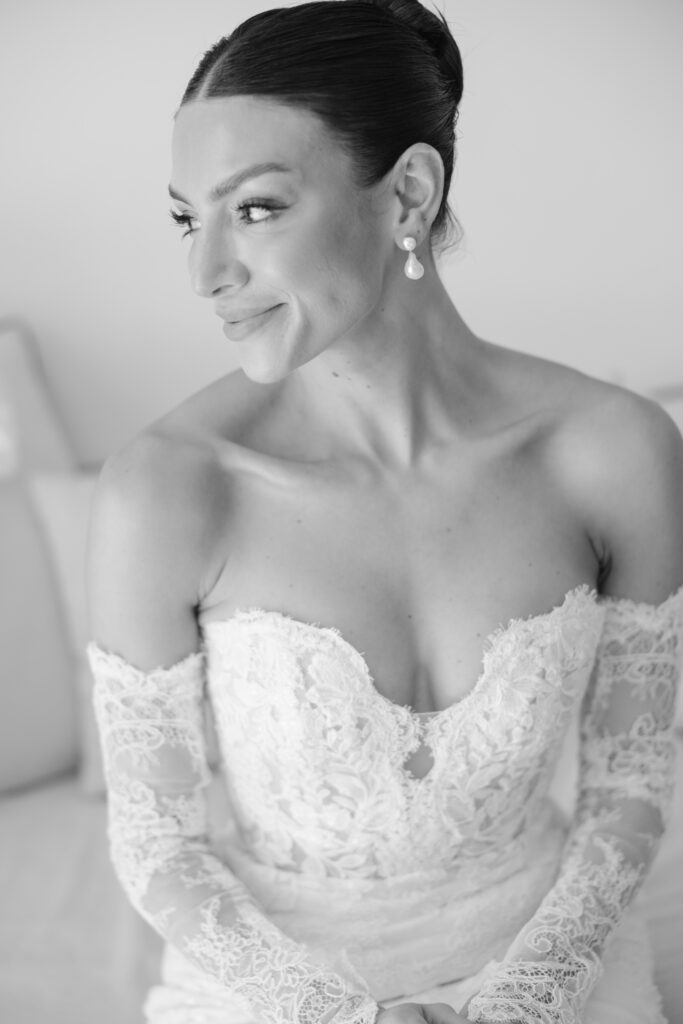 A woman in an off-the-shoulder lace wedding dress with long sleeves sits, smiling and looking to the side. She has her hair styled in a sleek bun and is wearing drop earrings. The photo is black and white.