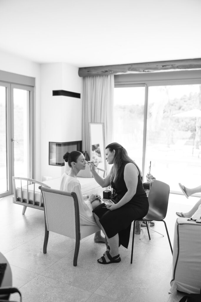 A makeup artist applies makeup to a seated woman in a bright room with large windows, a mirror, and modern furniture. The atmosphere is calm and focused.