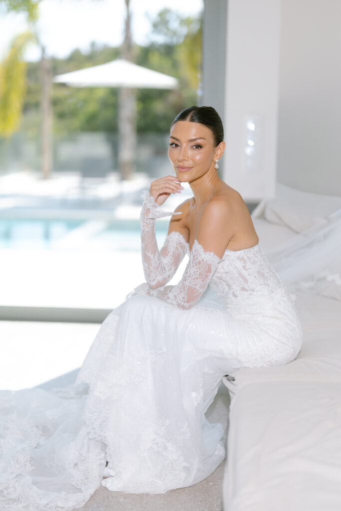 A bride in an off-the-shoulder lace wedding dress and gloves sits on a white sofa, smiling softly, with a sunlit pool and palm trees visible through a large window behind her.