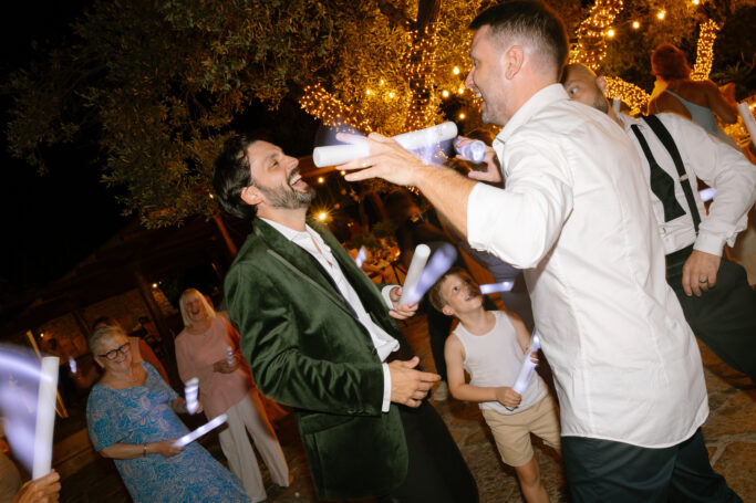 A group of people dance and celebrate outdoors at night under string lights. Two men in the foreground laugh and hold glowing sticks, while others join the festivities in the background, all dressed in semi-formal attire.