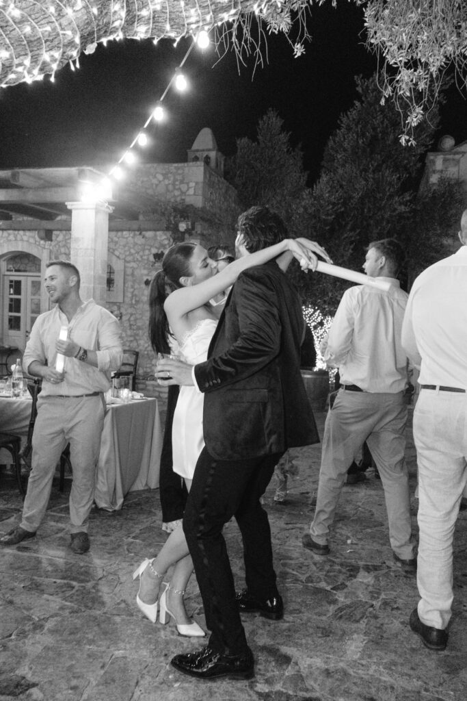 A couple dances closely outdoors at night under string lights, surrounded by other people mingling and dancing at a festive, possibly wedding, celebration. The scene is lively and joyful.