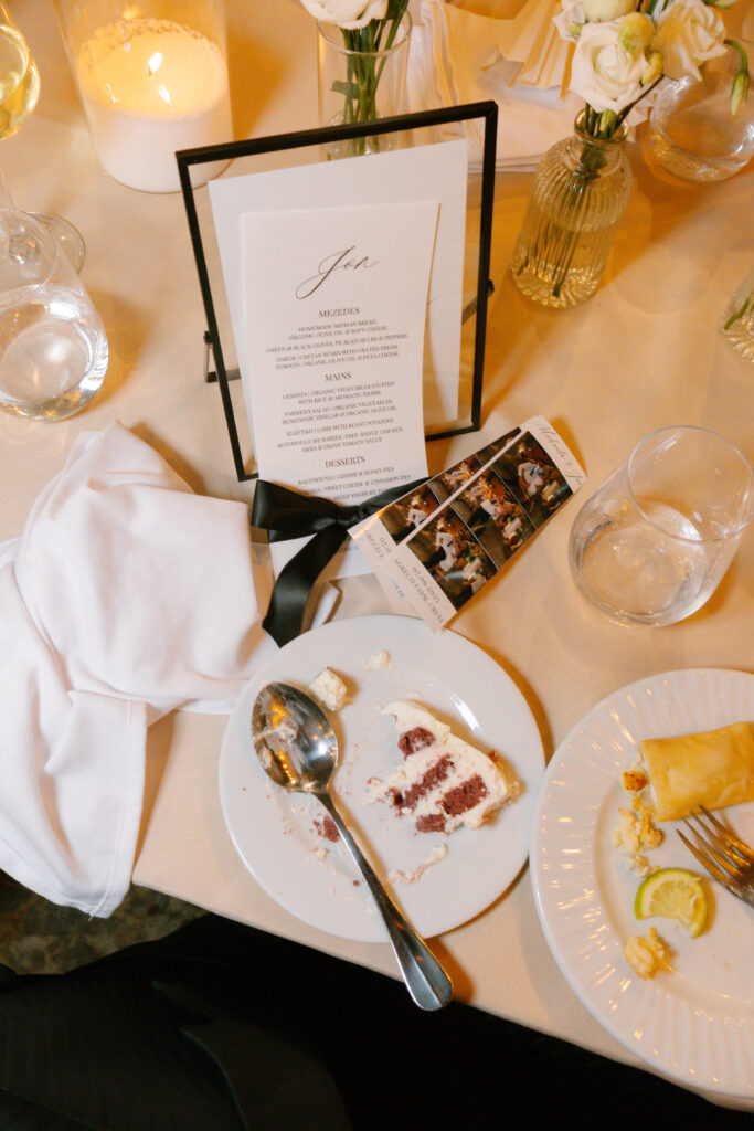 A table setting with a white napkin, two plates with partially eaten desserts, a menu in a black frame, a candle, flowers in vases, and a photo strip lying beside the plates.