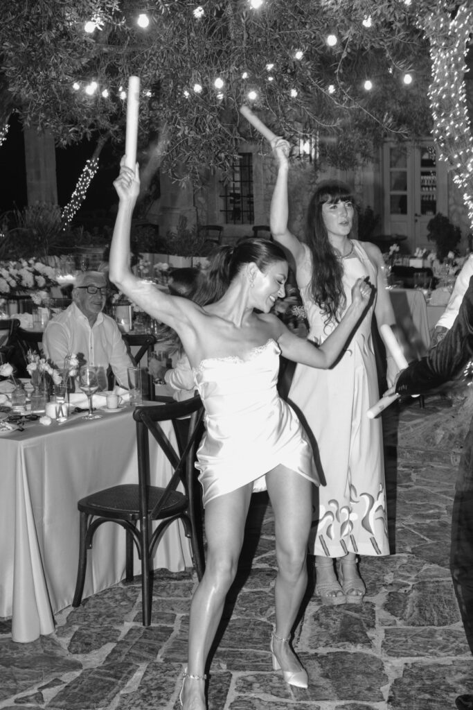 Two women dance energetically under string lights at an outdoor party, holding glow sticks. People sit at nearby tables, enjoying food and drinks. The scene is festive and joyful, captured in black and white.