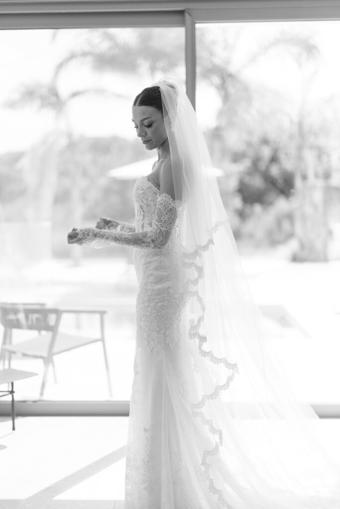 A bride in a lace wedding dress and long veil stands indoors by a large window, softly lit by natural light, looking down at her hands with a serene expression.