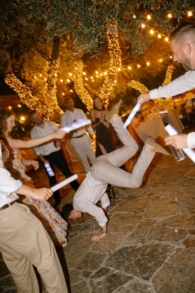 A person in light-colored clothes does a handstand on a stone patio at an outdoor party while others in festive attire cheer and wave glow sticks under trees decorated with string lights.