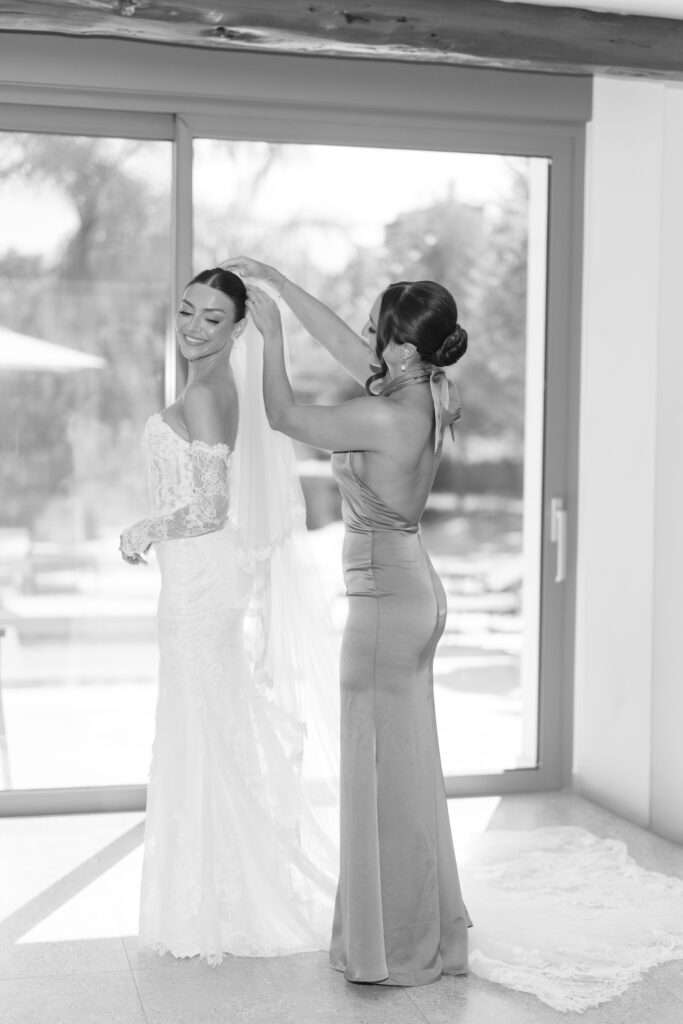 A bride in a lace wedding dress smiles as another woman in a fitted gown adjusts her veil. They stand indoors by large glass doors with sunlight streaming in.