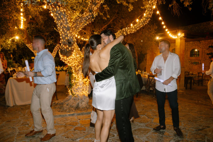A couple in formal attire kisses under string lights at an outdoor evening event, surrounded by people dancing and socializing near a large tree wrapped in fairy lights.