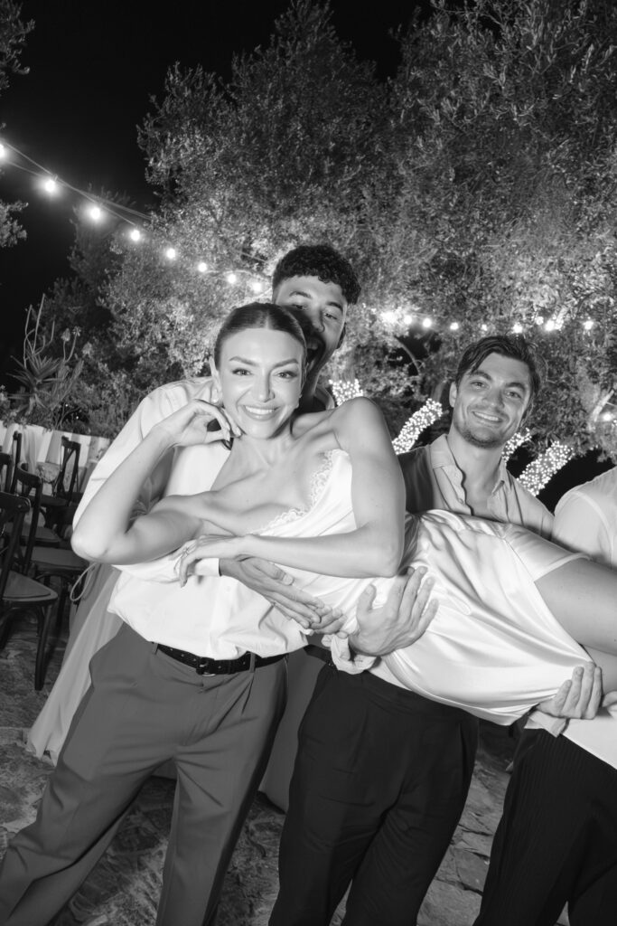 Three people smile as they hold up a fourth person in a playful pose at an outdoor nighttime event with string lights and trees in the background. The scene is captured in black and white.