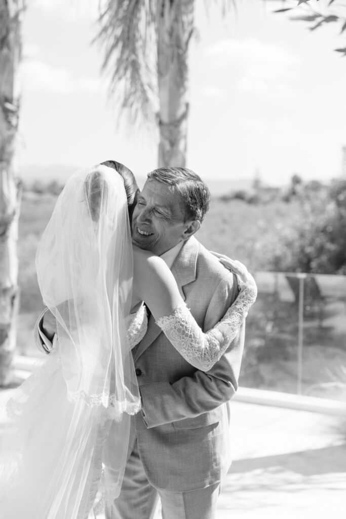 A bride in a lace wedding dress and veil hugs an older man in a suit outdoors. They both smile warmly, sharing a joyful moment. Palm trees and a glass railing are visible in the background. The image is in black and white.