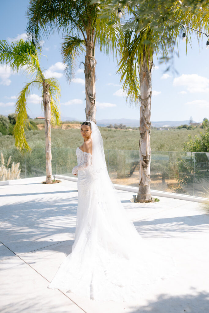A bride in a long white dress and veil stands outdoors on a sunny day, surrounded by palm trees, with a scenic background of hills, greenery, and a glass railing.