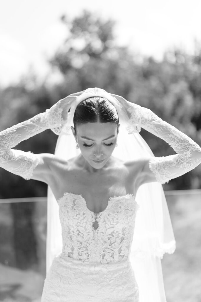A bride in a lace wedding dress and long lace gloves adjusts her veil outdoors. She has her eyes closed and head slightly bowed, with greenery blurred in the background. Black and white photo.